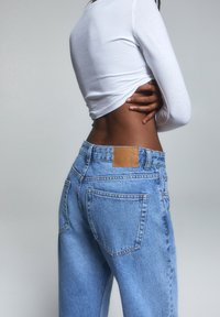 Jeans de mezclilla azul claro con cintura alta, con un parche de cuero marrón en la parte posterior y bolsillos estándar, combinados con una blusa blanca de manga larga.