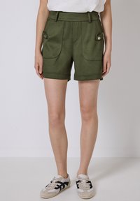 Žena oblečená v olivově zelených šortkách s předními kapsami a bílých teniskách s černými pruhy, stojící na jednobarevném pozadí.