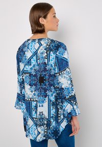 Blaue und weiße gemusterte Tunika mit ausgestellten Ärmeln, die florale und geometrische Designs in verschiedenen Blautönen auf einem hellen Hintergrund zeigt.