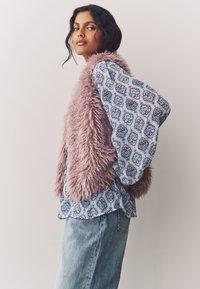 Faux fur vest in soft pink over a patterned blue top with flowing sleeves, paired with light denim jeans. Simple design, textured materials.