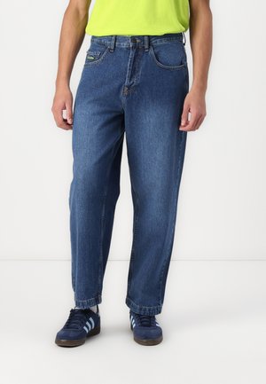 Denim jeans in a dark blue color, featuring a relaxed fit, five pockets, and a button closure. Worn with dark blue sneakers.