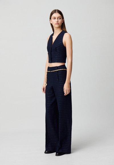 Gilet navy court avec un col en V et des boutons sur le devant, associé à un pantalon large avec une ceinture en chaîne dorée et une texture à rayures subtile.
