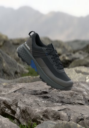 Chaussure de randonnée Columbia noire et grise avec des accents bleus flottant au-dessus d'un terrain rocheux dans un paysage de montagne en plein air.