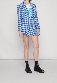 Blazer en estampado houndstooth azul y blanco con una falda mini con volantes, combinado con una camiseta gráfica y botines negros con detalles dorados.