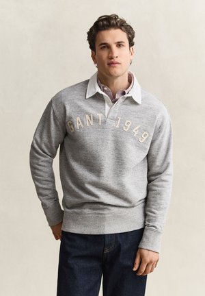 Jonge man draagt een grijze GANT 1949 sweater over een wit hemd met kraag en donkerblauwe spijkerbroek, staand tegen een neutrale achtergrond.