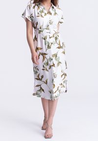 Donna che indossa un vestito midi bianco con stampe floreali verdi e marroni, una cintura abbinata e sandali con tacco alto beige.