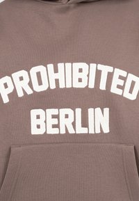 Felpa marrone con testo bianco testurizzato "PROHIBITED BERLIN" sopra una tasca frontale.