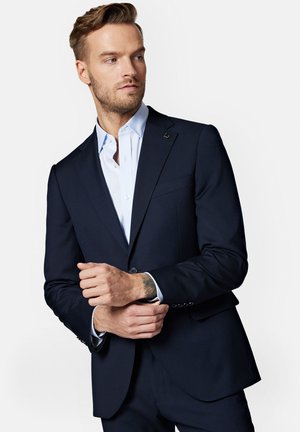 Granatowy garnitur o dopasowanym kroju, z ostrym klapą, zapinany na jeden guzik, z kontrastową jasnoniebieską koszulą pod spodem.