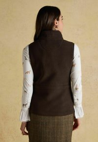 Braune Fleece-Weste mit hohem Kragen, mit Stickerei auf der Rückseite. Getragen über einem hellen gemusterten Hemd und einem karierten Rock. Strukturiertes Material.