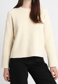 Rippiger cremefarbener Pullover mit Rundhalsausschnitt und breiten Bündchen, der eine lässige Passform und strukturiertes Material aufweist, kombiniert mit schwarzen Jeans.