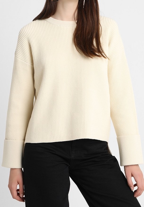 Rippiger cremefarbener Pullover mit Rundhalsausschnitt und breiten Bündchen, der eine lässige Passform und strukturiertes Material aufweist, kombiniert mit schwarzen Jeans.