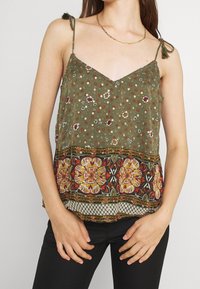 Olivgrön ärmlös topp med blommönster, med spetsdetalj vid nederkanten och färgglada geometriska inslag i botten.