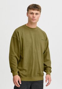 Oliwkowy sweter z okrągłym dekoltem, obniżonymi ramionami i ściślejszymi mankietami. Posiada minimalne logo na piersi i miękką teksturę.