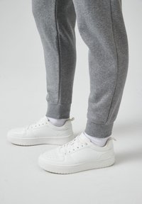 Weiße Sneaker mit einer glatten Lederoberfläche, ausgestattet mit durchbrochenen Details, kombiniert mit grauen Jogginghosen, die gerippte Bündchen haben.