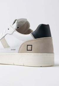 Sneaker bianco con dettagli in suede beige, tallone nero e suola testurizzata. Presenta un logo quadrato e un branding sottile sul lato.
