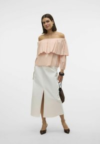 Top rosa off-the-shoulder con rouches sovrapposti, abbinato a una gonna midi bianca con uno spacco frontale e accessori marroni.