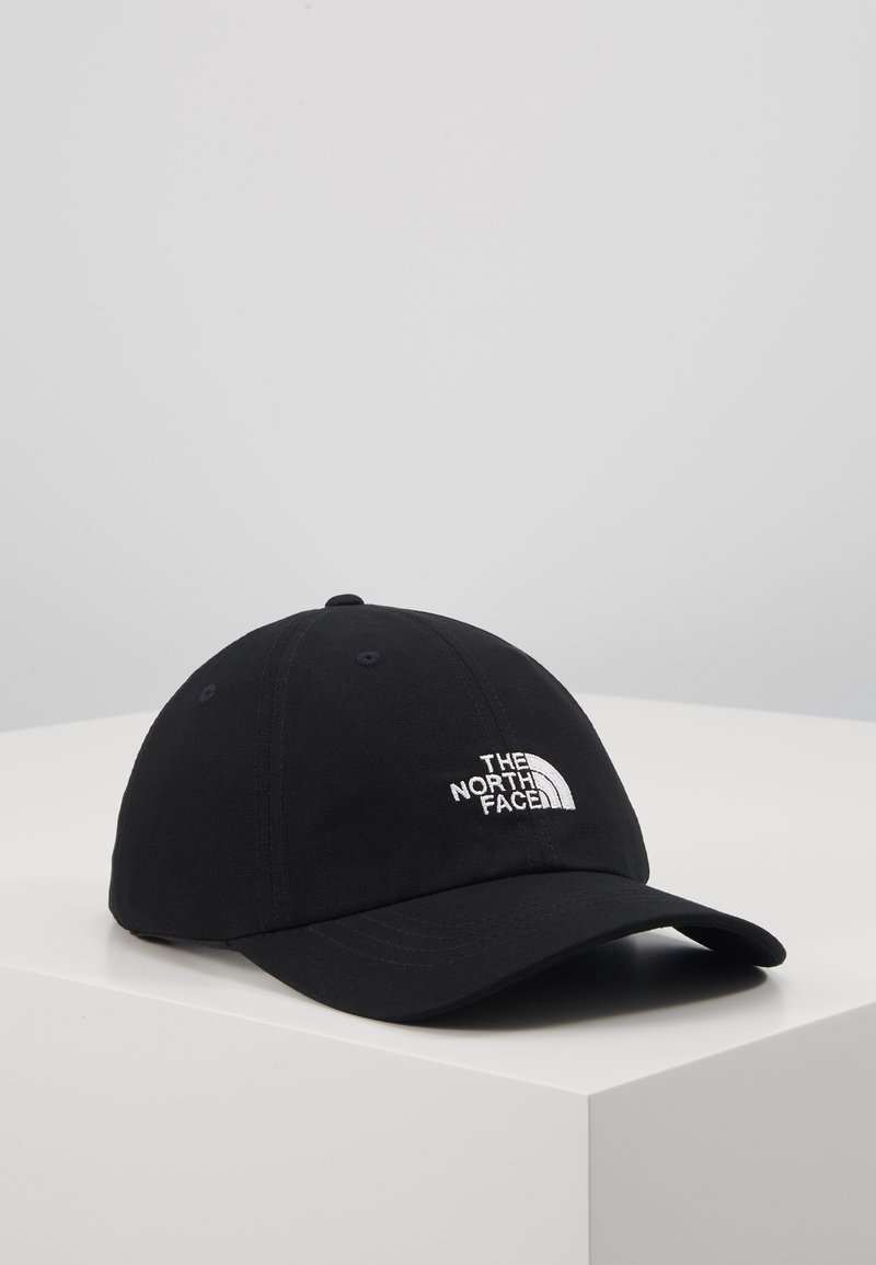 Casquette de baseball noire à visière incurvée avec logo brodé blanc "The North Face", posée sur une surface blanche plane sur fond gris clair.