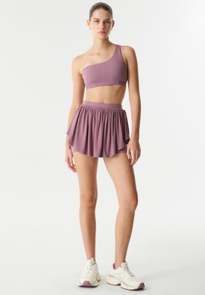 Reggiseno sportivo lavanda a spalla unica abbinato a una gonna plissettata, entrambi in tessuto leggero. Indossato con scarpe da ginnastica bianche e chunky.