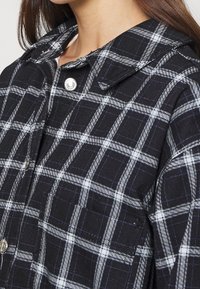 Närbild av en person som bär en svart-och-vit rutigt skjorta med knappar och spetskrage samt silverfärgade knappar.