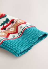 Gros plan d'un poignet de manche tricotée présentant un tissu côtelé bleu sarcelle et un motif coloré avec des arbres, des zigzags et des formes losanges en rouge, beige et vert.