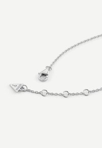 Srebrna bransoletka łańcuszkowa z owalnymi ogniwami, okrągłymi zdobieniami oraz trójkątną zawieszką z napisem "Guess". Zapięcie na klamrę w komplecie.