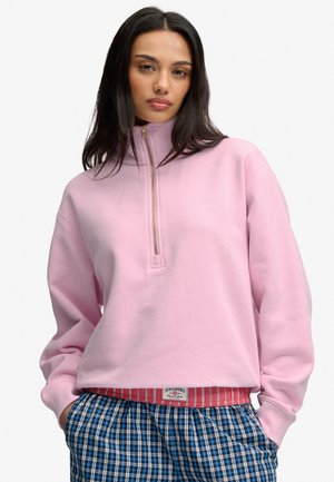 Donna con lunghi capelli scuri che indossa una felpa rosa con zip a quarto e pantaloni blu a quadri, in piedi con le mani nelle tasche su sfondo bianco.