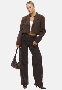 Blazer marron court sur un top blanc, associé à un jean ample marron foncé. Les accessoires comprennent un sac marron et un collier en perles.