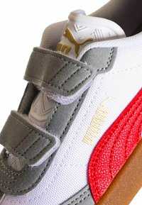 Sneaker bianca con accenti grigi e rossi, materiali testurizzati, fascette in velcro, un logo dorato e una suola in gomma naturale.