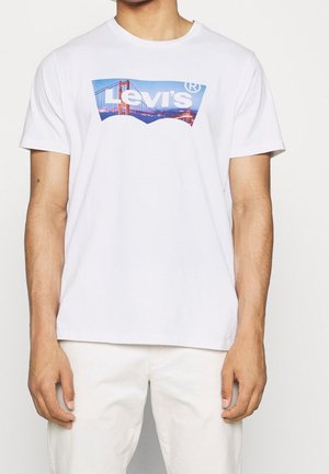 Mężczyzna ubrany w białą koszulkę Levi's z grafiką Mostu Golden Gate i białe spodnie, stojący na jednolitym tle.