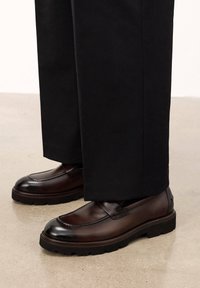 Mocasines de cuero marrón con acabado brillante, suelas de goma y parte superior cosida, combinados con pantalones negros ligeramente holgados.