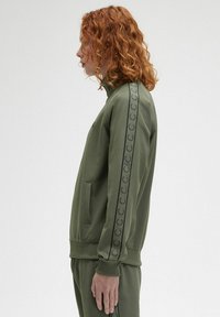 Felpa verde oliva con zip e collo alto, dotata di stampa del logo nera lungo le maniche e tasche laterali. Tessuto morbido e testurizzato.