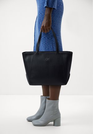 Black leather tote bag with embossed 'CK' logo, featuring dual top handles, structured shape, and subtle side accents. Light gray ankle boots with block heels.
