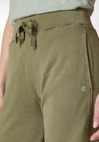 Olijfgroene broek met trekkoord, gemaakt van zachte, gestructureerde stof. De broek heeft een zijzak en een klein logo-detaillering nabij de tailleband.