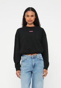 Sweatshirt scurt negru cu mâneci lungi, având un logo roz pe piept. Asortat cu blugi din denim de culoare albastru deschis.