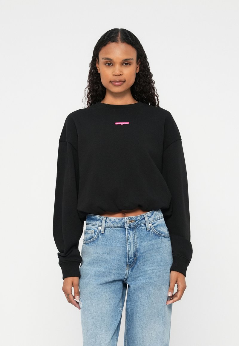 Sweatshirt scurt negru cu mâneci lungi, având un logo roz pe piept. Asortat cu blugi din denim de culoare albastru deschis.