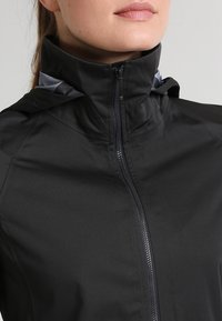 Mujer vistiendo una chaqueta negra con cremallera y un alto cuello, con un vistazo de una capucha gris debajo, contra un fondo neutro.