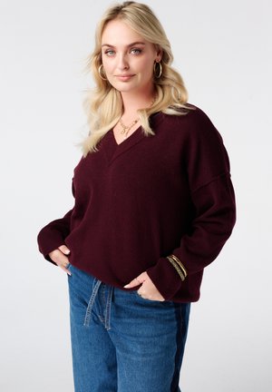 Blonde vrouw in een bordeauxrode V-hals trui en blauwe spijkerbroek, draagt gouden creolen, ketting en armbanden, handen in de zakken, tegen een witte achtergrond.