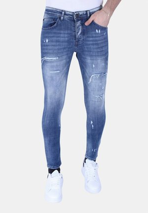 Man draagt slim-fit versleten blauwe spijkerbroek met scheuren en witte sneakers, hand in zak, tegen een effen achtergrond.