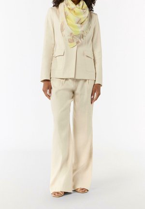 Femme portant un costume beige avec un pantalon à jambes larges, un blazer assorti, une écharpe jaune pastel à motifs et des talons ouverts beiges, debout devant un fond blanc.