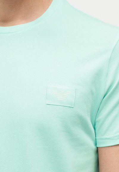 Κοντινό πλάνο ενός ατόμου που φοράει ένα ανοιχτό τιρκουάζ μπλουζάκι με στρογγυλή λαιμόκοψη και ένα μικρό, τονικό μπάλωμα με λογότυπο αετού στο στήθος.