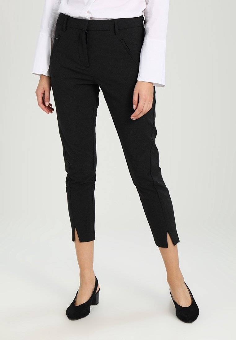 Femme portant un pantalon slim noir aux chevilles avec des fentes devant, une chemise blanche à manches longues et des chaussures slingback noires à petit talon bloc.