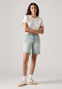 Ανοιχτό μπλε τζιν σορτσάκι με ψηλή μέση, διπλωμένη γωνία και δύο μπροστινές τσέπες, συνδυασμένο με κρεμ κοντομάνικο μπλουζάκι και αθλητικά παπούτσια.