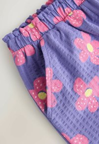 Közelkép lila texturált szövetből készült rövidnadrágról, rózsaszín és sárga virágmintákkal és elasztikus derékpánttal.