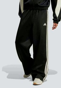 Fekete Adidas tréningnadrágot viselő személy, amelyen fehér csíkok találhatók, bézs sportcipőben áll, az egyik lábát kissé előre helyezve.