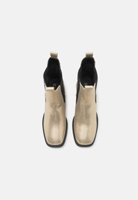 Bottines dorées avec une finition métallique texturée, panneaux latéraux élastiques noirs et languette pour un enfilage facile. Semelle noire plate.