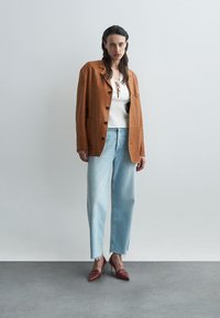 Sacou din piele de căprioară maro cu nasturi mari, bluză albă cu textură, blugi deschiși cu aspect uzat în nuanță de albastru și pantofi cu toc înalt roșii, așezate pe o suprafață gri.