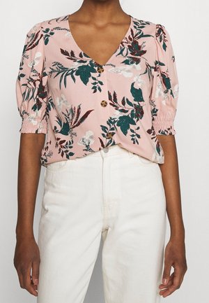 Kvinde iført lyserød kortærmet bluse med grøn og bordeaux botanisk print og beige højttaljede bukser.