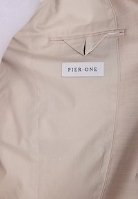 Intérieur en tissu beige avec une poche et une étiquette indiquant "PIER•ONE" en texte noir. Texture lisse avec un léger éclat.