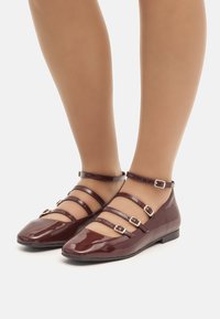 Ballerines en cuir verni bordeaux avec un bout arrondi, plusieurs fines lanières avec des boucles dorées, et un petit talon plat.