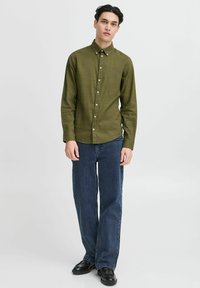 Olivenfarget skjorte med krage og subtil tekstur, kombinert med vide mørkeblå jeans og sorte sko.